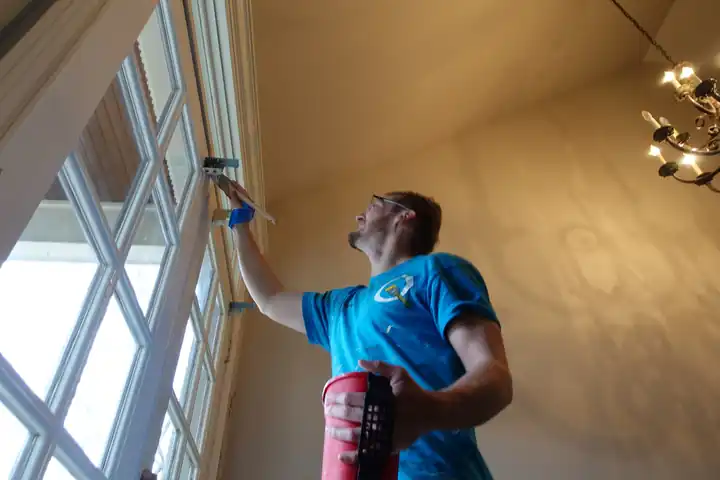 A painter brushing the grid of a tall arched window under a vaulted ceiling